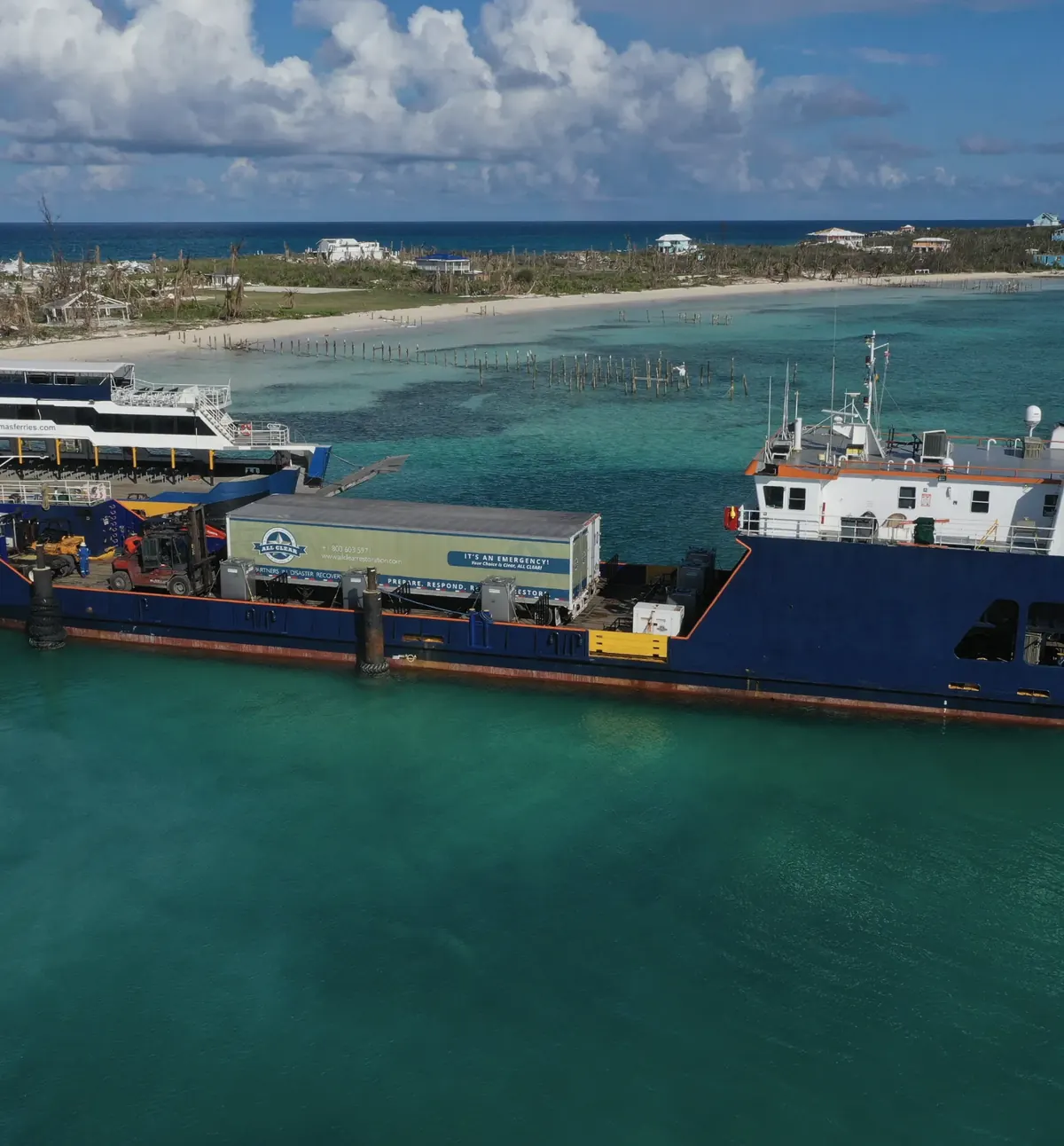 An ALL CLEAR equipment trailer on the deck of a supply ship, with a storm-stripped shoreline behind