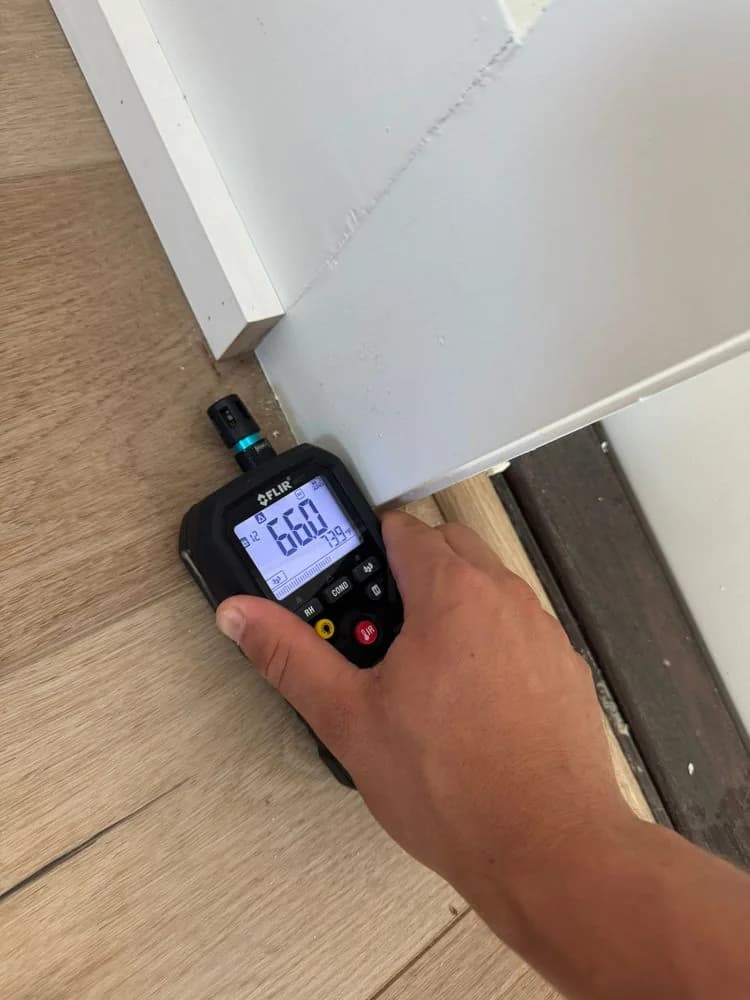 A moisture meter reading 66% on wood flooring beside a patio door