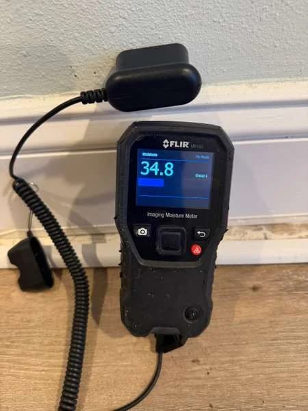 A moisture meter reading 34.8 on the drywall above the baseboard