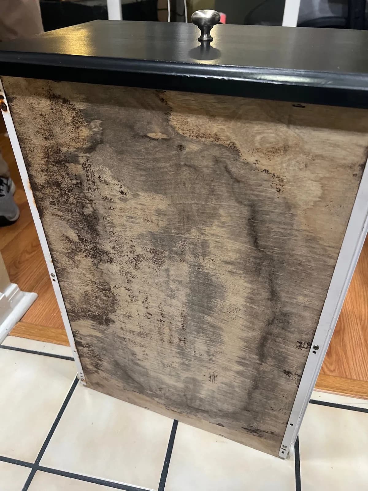 A removed cabinet panel standing on a tile floor. The wood is mottled with brown and black water stains and mold.