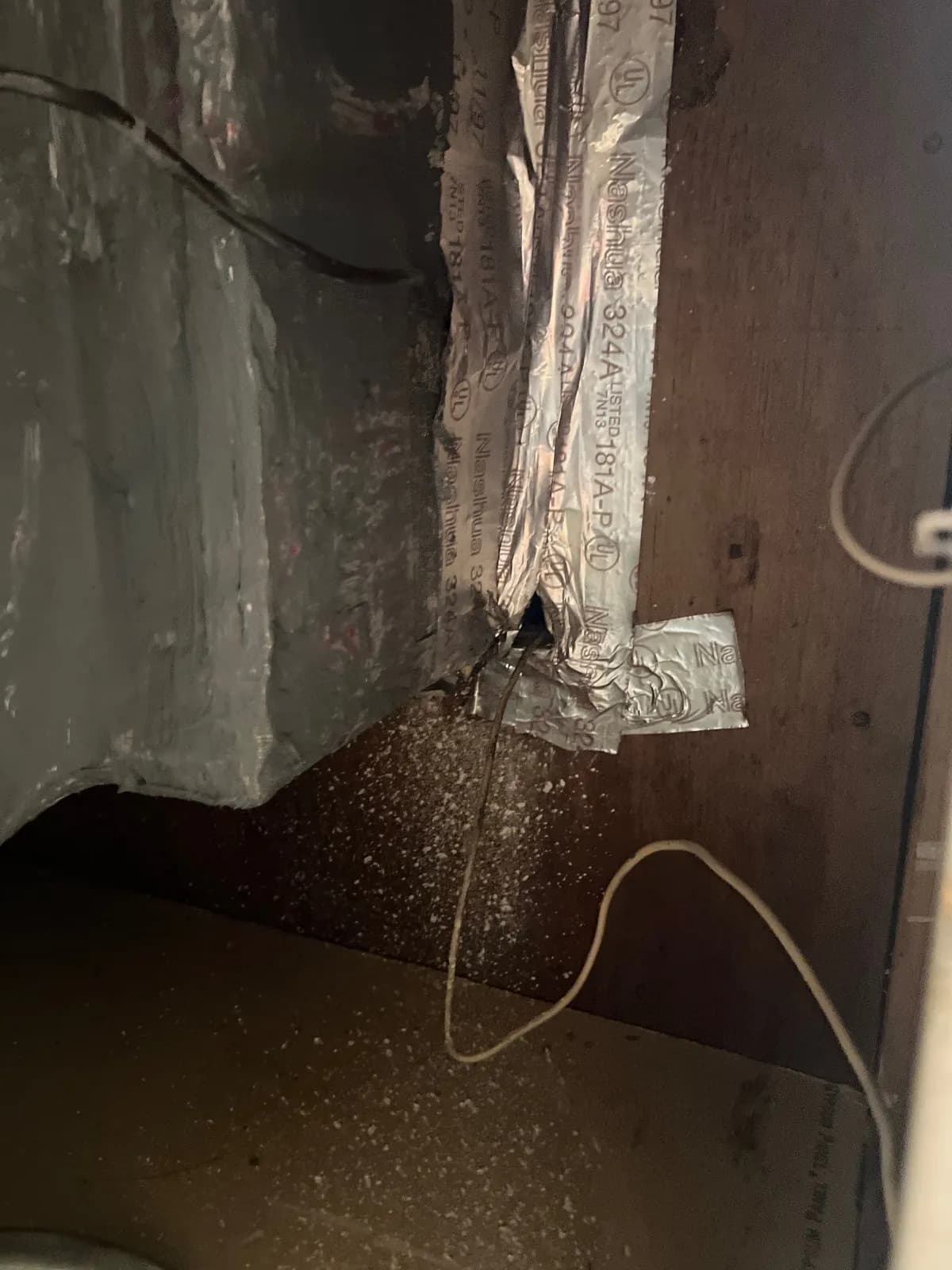 Dim view inside an air handler closet showing foil-wrapped ductwork, a hanging wire and debris scattered on a dark surface below.
