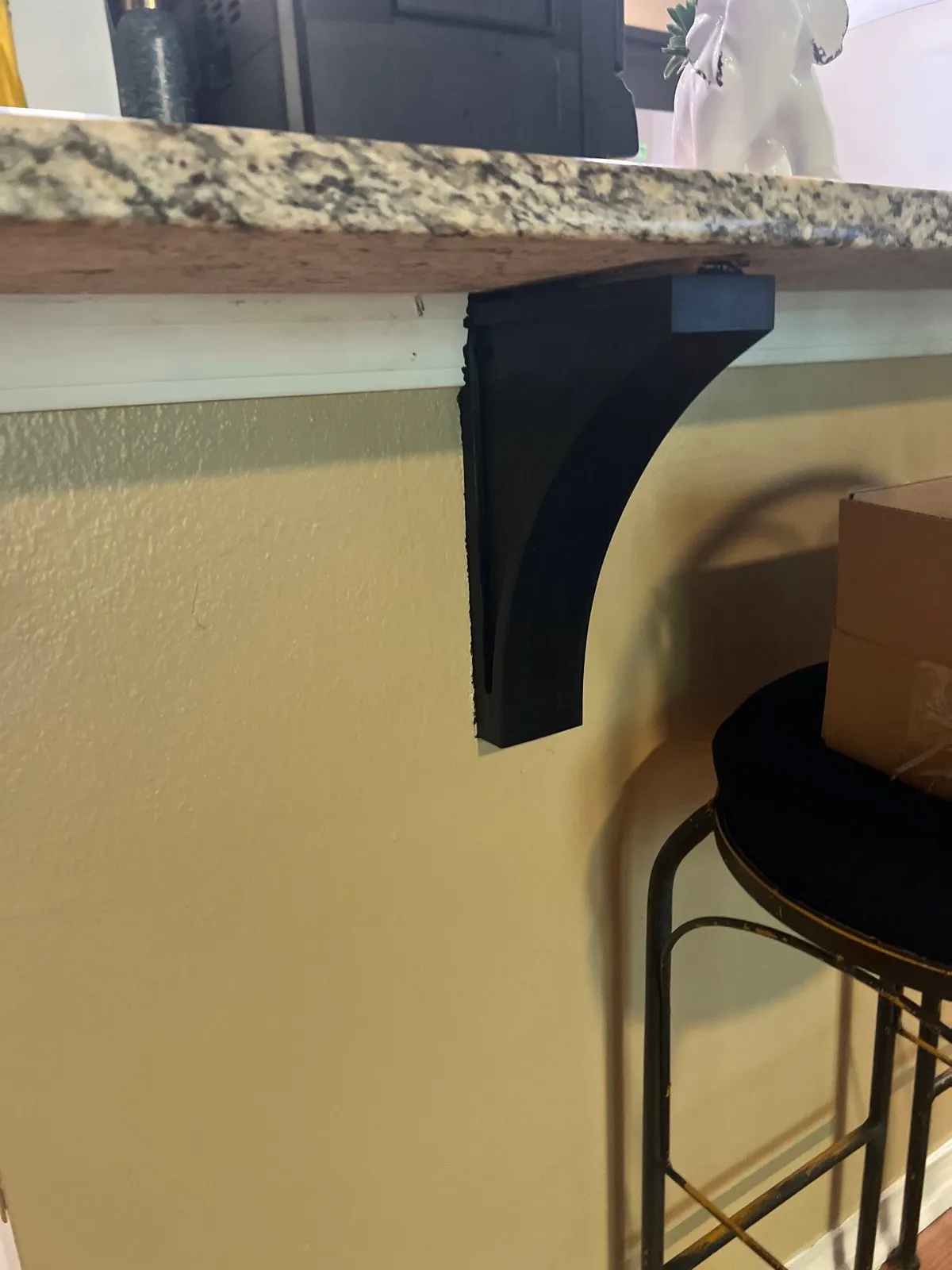 The side of a kitchen bar with a granite top, a black support bracket and a beige painted wall, with a bar stool next to it.