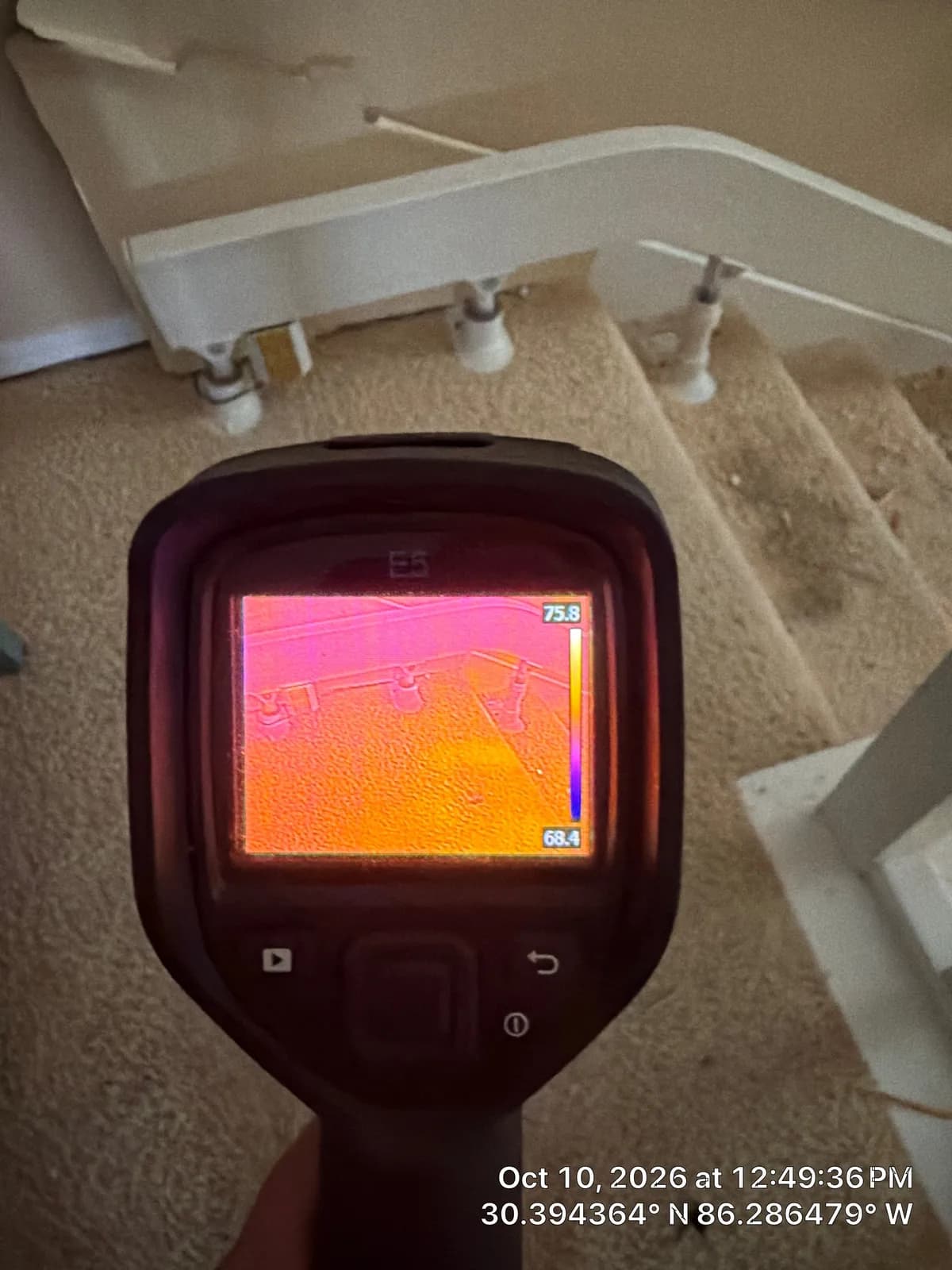 Hand holding a thermal camera. Its screen shows an orange and pink heat image of the stair railing and wall.