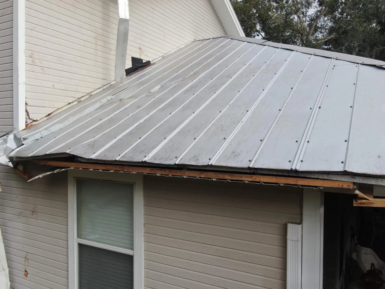 Gray ribbed metal roof over light siding. The panels are wavy and bent, and the trim along the lower edge is broken.