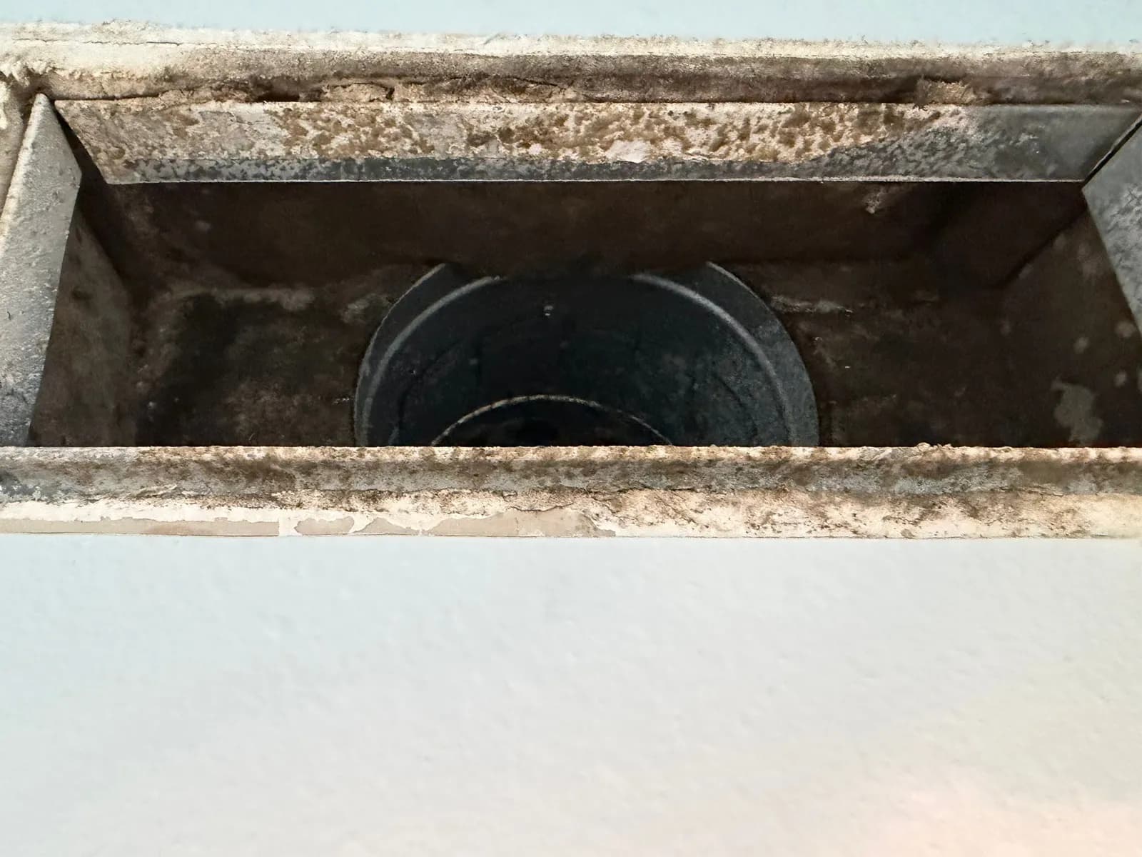 Rectangular metal vent box in a wall with dark specks and staining around its edges and a round duct connection inside.