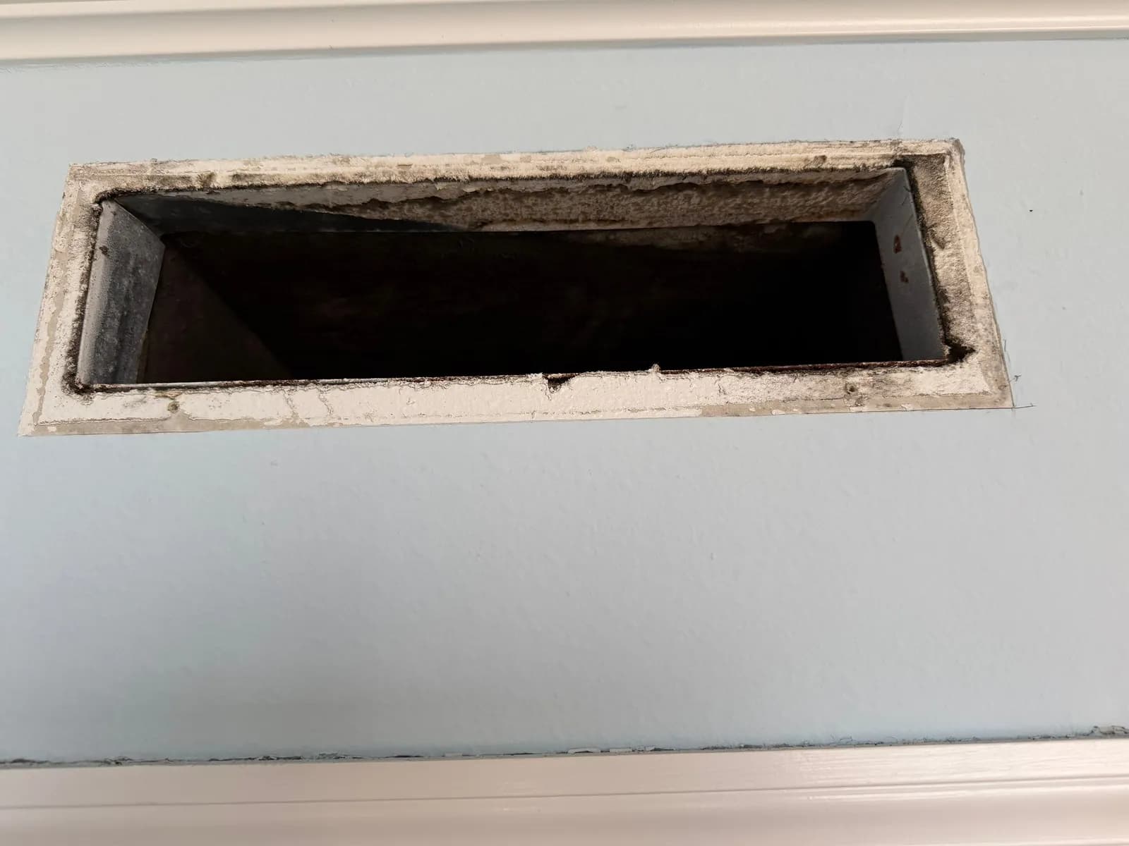 Rectangular wall opening for a vent, edges coated in dark and white residue, light blue wall around it.