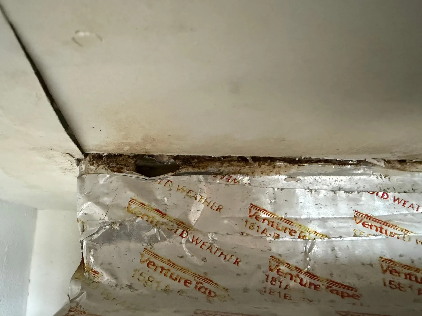Foil tape with printed red lettering along a duct seam, with dark mold spots where it meets a white surface.