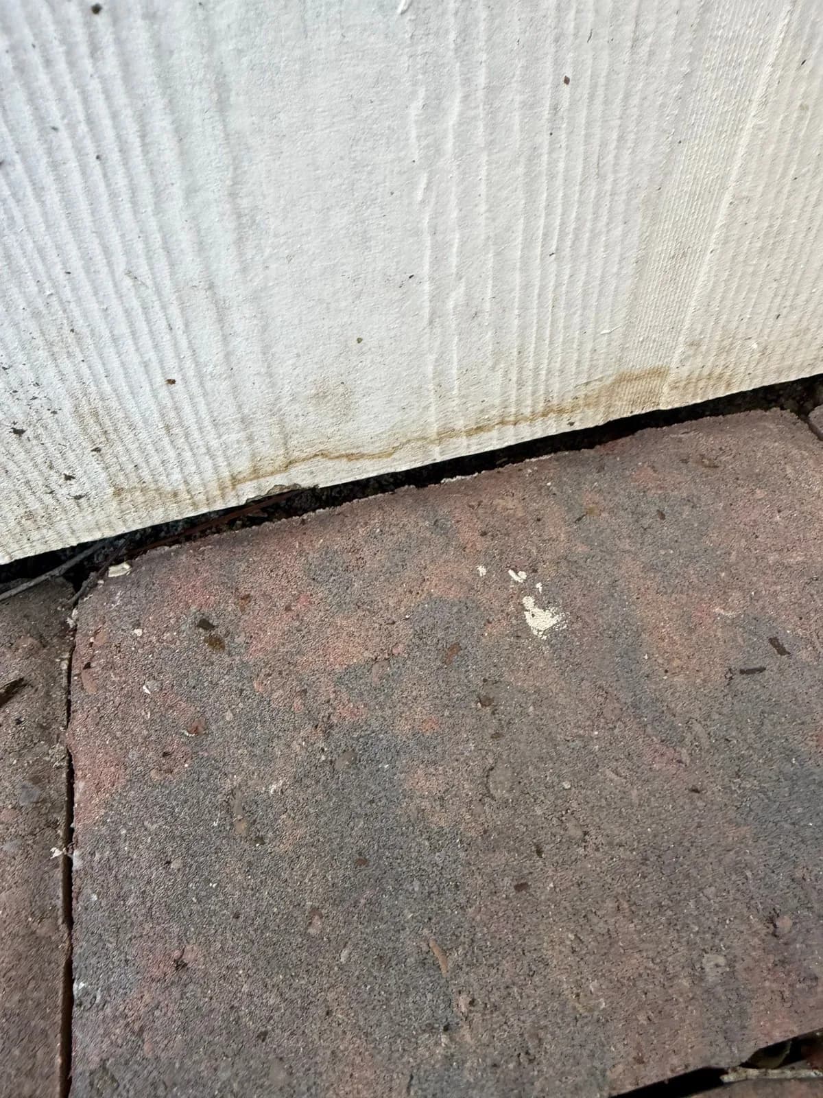 Close-up of white textured siding meeting a reddish brick paver, with a brown stain line along the bottom of the siding.