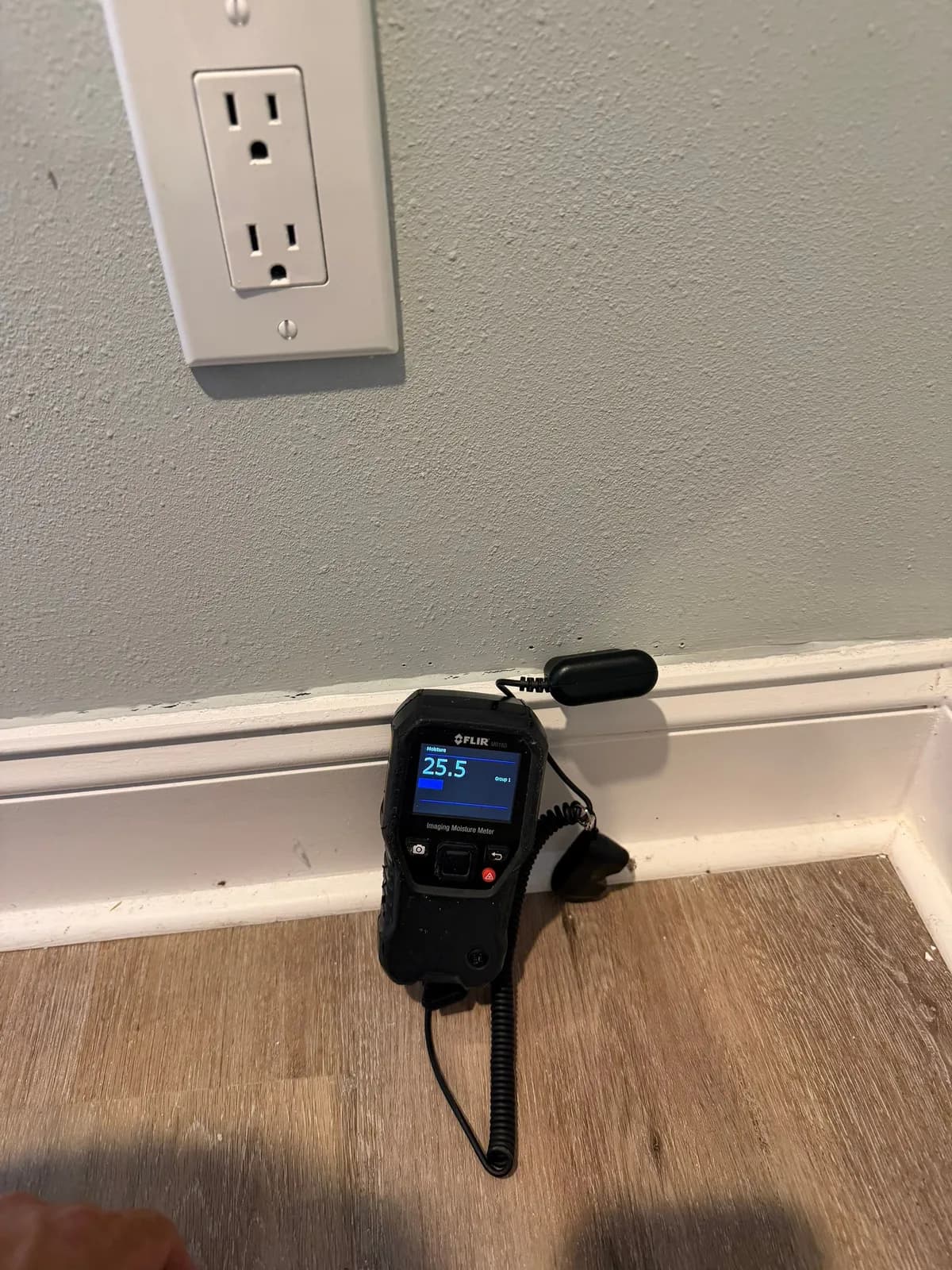 A black handheld moisture meter showing 25.5 held against a gray-green wall below an outlet, above white baseboard and wood-look flooring.