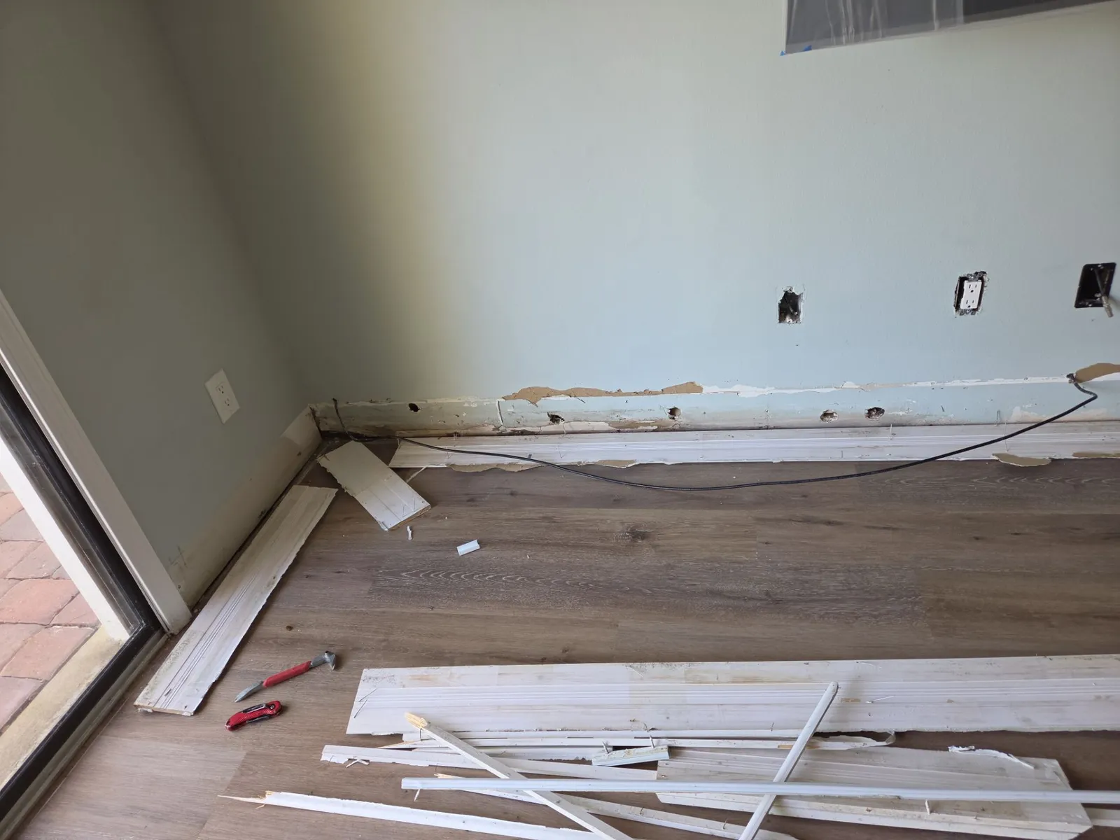 Living room wall with baseboards pulled off and stacked on wood-look flooring, a sliding glass door at the left edge.