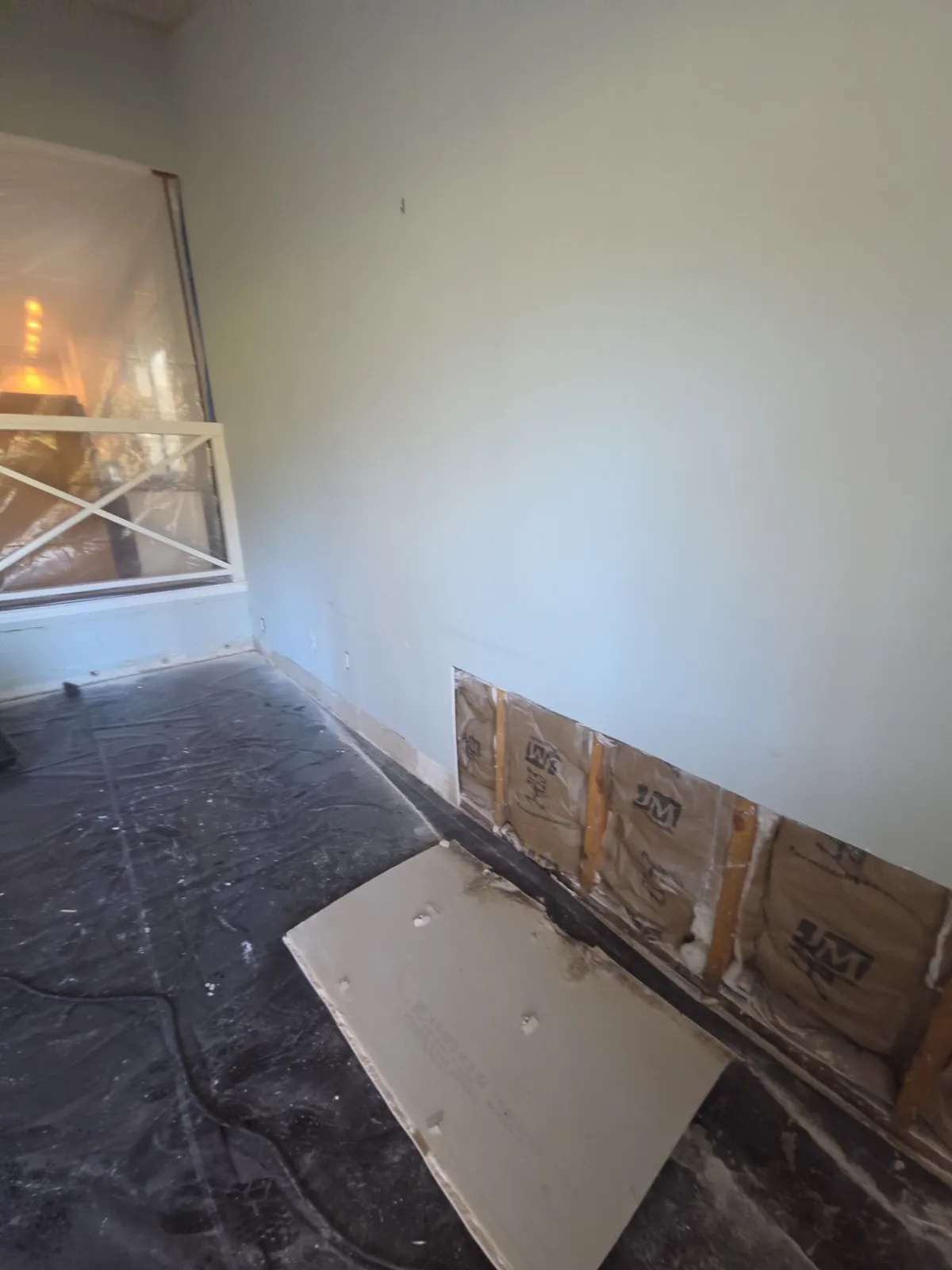 Room with plastic on the floor and the bottom strip of drywall removed along one wall, exposing paper-faced insulation between studs.