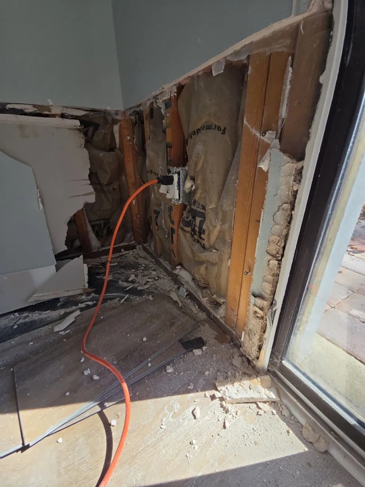 Lower wall next to a sliding glass door cut open to show wood studs, insulation and an outlet box, with debris on the floor and an orange cord.