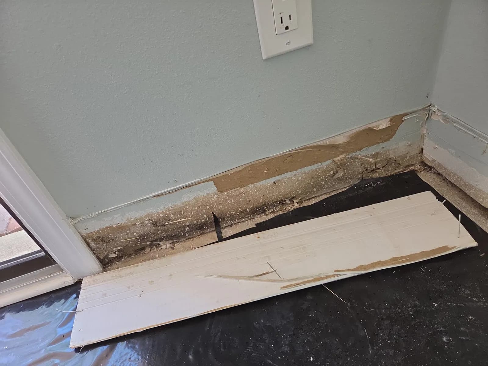 Bottom of a wall beside a sliding door with baseboard removed, showing dark stained drywall along the floor line and a loose board on the floor.
