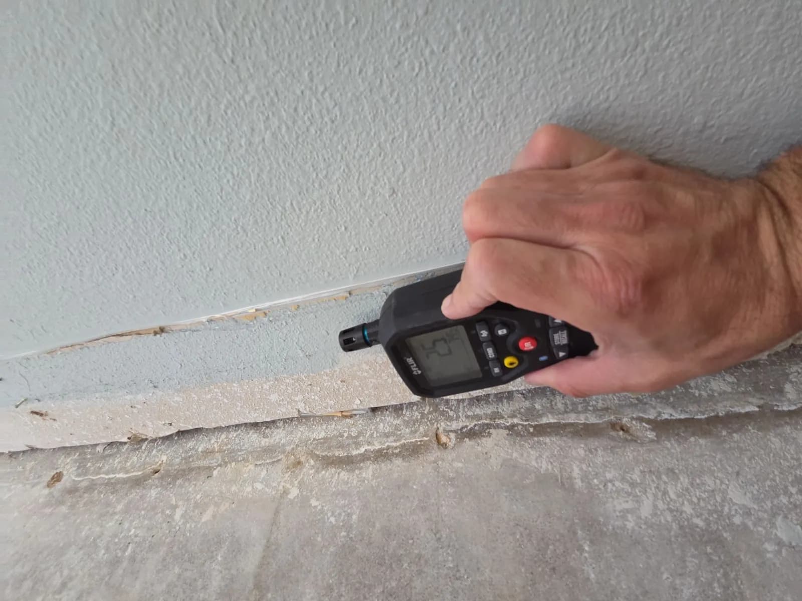 A hand holding a pin-type moisture meter against a gray wall just above where the lower drywall was cut away.